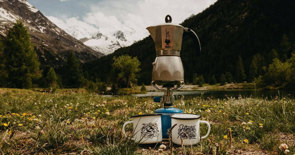 Stovetop moka pot brewing on a camping burner in a mountain meadow with two enamel mugs