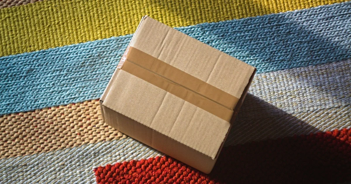 A plain taped cardboard parcel with no seller markings resting on a striped rug, the unlabeled delivery at the end of any top Amazon gadgets 2026 purchase