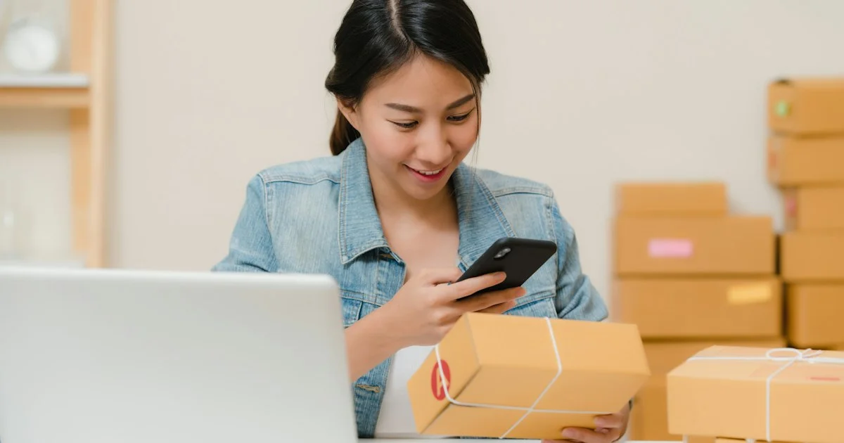 Small-business owner checking a parcel against her phone beside stacked shipping boxes, the everyday job a top barcode scanner app 2026 buyers shortlist has to do