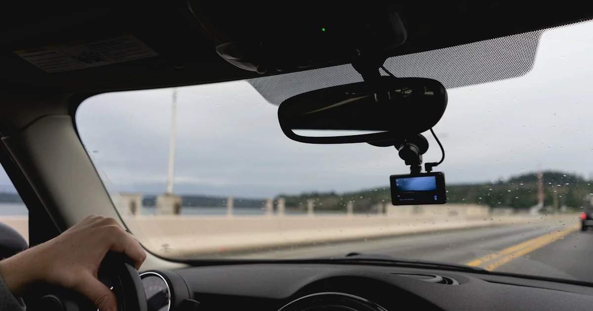 A small dash cam mounted behind the rear-view mirror while the driver holds the wheel on a bridge, the mounting position to plan for when deciding which dash cam to buy 2026
