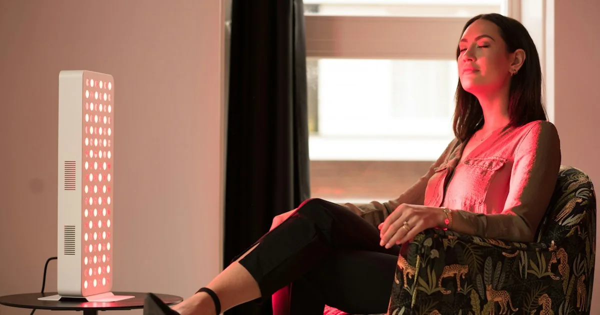 A woman seated several feet from a red LED panel on a side table, the treatment distance that decides which red light therapy device should I buy